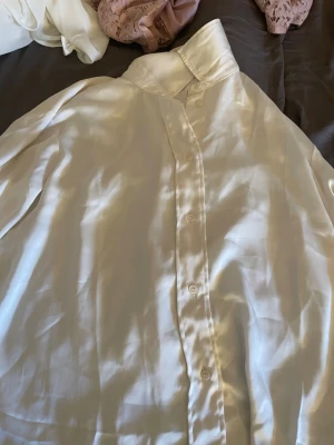 Vit sidenliknande skjorta med knappar - Snygg vit skjorta i glansigt material med klassisk krage och knäppning framtill. Skjortan har långa ärmar och en clean, minimalistisk look som passar till många olika stilar. Perfekt för dig som gillar en stilren vibe.