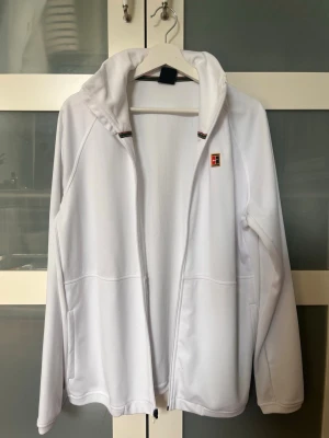 Vit Nike Dri-FIT zipup - Snygg vit hoodie från Nike med Dri-FIT-material som andas. Jackan har dragkedja, huva och två sidofickor. Liten logga på bröstet och sportig design, perfekt för träning eller chill.