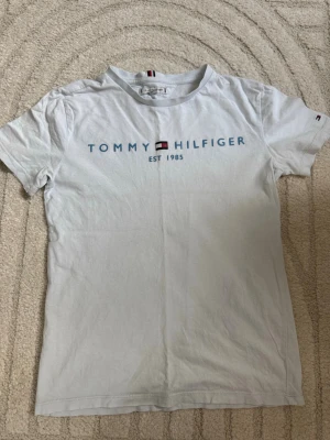 Vit Tommy Hilfiger t-shirt - Vit t-shirt från Tommy Hilfiger med blå logotyp och text 'EST 1985' framtill. Klassisk passform med korta ärmar och rund hals.  Hilfiger-detaljer på ärmen och i nacken. 