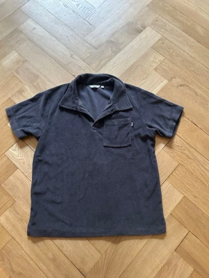 Mörkgrå frotté piké  - Mjuk och skön mörkgrå t-shirt i frotté med korta ärmar och en avslappnad krage. Bröstficka på vänster sida för extra stilpoäng. Perfekt för dig som gillar en chill och unik look.
