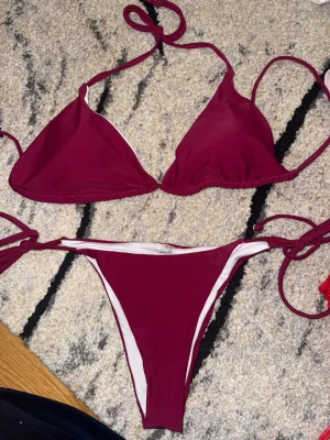 Vinröd bikini med knyt  - Nyskick. Snygg vinröd bikini med vita detaljer och knyt i sidorna samt runt nacken. Klassisk trekantsmodell upptill och smala band. Tillverkad i stretchigt material som sitter bekvämt på kroppen. Perfekt för sommardagar på stranden eller vid poolen.
