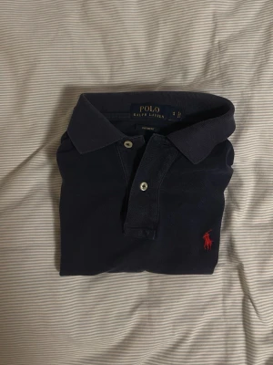 Mörkblå pikétröja från Ralph Lauren - Klassisk mörkblå pikétröja från Ralph Lauren med röd broderad logga på bröstet. Tröjan har krage, knappar framtill och är i en custom fit-modell. Perfekt för en stilren och avslappnad look.