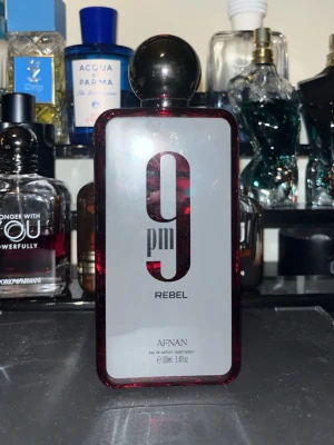Afnan 9PM Rebel Eau de Parfum 100ml - Afnan 9PM Rebel är en parfym i en rektangulär flaska med mörkröd ram och stor svart rund kork. Flaskan har en modern design med tydlig logotyp och text i svart. Innehåller 100 ml eau de parfum som passar dig som gillar unika och intensiva dofter. 80/100 ml