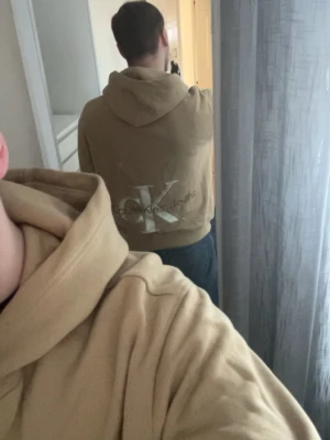 Beige hoodie från Calvin Klein Jeans - Snygg beige hoodie från Calvin Klein Jeans med stor logga på ryggen och liten CK-logga framtill. Klassisk känguruficka och huva. Perfekt för en avslappnad och trendig stil. Mjuk bomullskänsla och ribbade muddar vid ärmslut och nederkant.