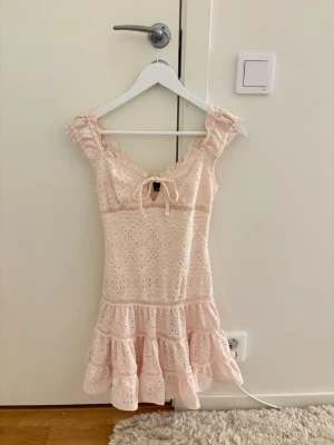 Ljusrosa spetsklänning med volang - Söt ljusrosa kortklänning med broderad spets och volang nertill. Klänningen har breda axelband med rynkade detaljer och en liten knytning framtill. Perfekt för sommarens alla tillfällen.