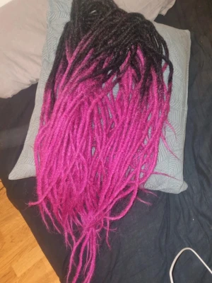 Svart och rosa syntetiska dreads - Snygga syntetiska dreads i ombre-stil, svart upptill som övergår i intensivt rosa. Perfekt för att skapa en unik look och enkelt att fästa i håret. Lätta och flexibla, passar dig som vill sticka ut med färg och stil. 110 cm från ände till ände, dessa är dubbeländade. 48 st, endast insatta några dagar så nyskick, dock har jag tvättat dom och några små gummiband kan sitta kvar. 