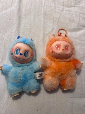 Två fluffiga monster-nyckelringar - Säljer två gulliga monster-nyckelringar från THE MONSTERS. Ena är blå med blå detaljer och den andra är orange med vita och orange detaljer. Båda har stora plastansikten med leende uttryck och fluffig päls. Perfekta som accessoar till väskan eller nycklarna.