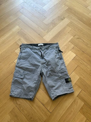  Stone Island shorts  - Säljer ett par grå cargo shorts från Stone Island med klassisk patch på benet. Shortsen har flera fickor, bälteshällor och en avslappnad passform. Perfekta för sommardagar och streetwear-stil.