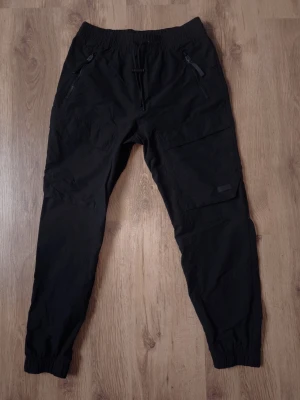 H&M Move Cargo Joggers – Herr – Bra skick -   Säljer ett par snygga och funktionella joggers från H&M Move. Perfekta för både träning, friluftsliv eller som en del av en avslappnad vardagsstil (techwear). Detaljer:  Märke: H&M Move.  Storlek: S (CN 175/80A).  Färg: Svart.  Material: Slitstarkt och stretchigt funktionsmaterial (94% polyamid, 6% elastan). Tyget är lätt, följsamt och torkar snabbt.