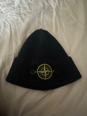 Svart mössa från Stone Island - Säljer en svart ribbstickad mössa från Stone Island med den klassiska loggan framtill i gult och grönt. Mössan är i mjukt material och har en uppvikt kant med tydlig patch. Perfekt för dig som gillar streetwear och vill ha en snygg accessoar.