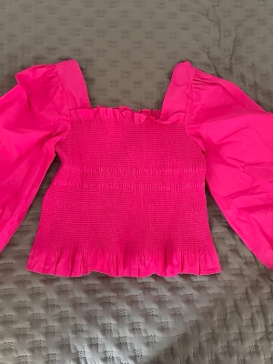 Rosa topp med puffärmar från NA-KD - Superfin rosa topp från NA-KD med smockad kropp och volangdetaljer. Toppen har långa puffärmar och en fyrkantig halsringning. Perfekt för dig som vill sticka ut med färg och trendiga detaljer.