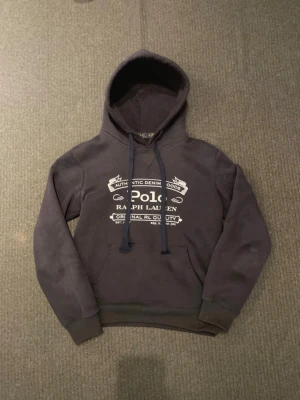 Marin blåhoodie från Polo Ralph Lauren - Snygg marinblå hoodie från Polo Ralph Lauren med vit tryckt logga och text på bröstet. Ser solblekt ut bilderna är ej de, något konstigt me ljuset bara 