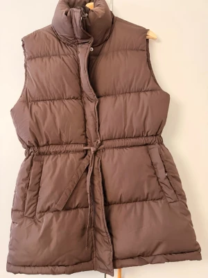 Brun dunväst från Bik Bok - Snygg brun dunväst från Bik Bok med hög krage och knytband i midjan. Västen har två stora fickor framtill och stängs med dragkedja och tryckknappar. Perfekt för lager på lager och ger en trendig look till din outfit.