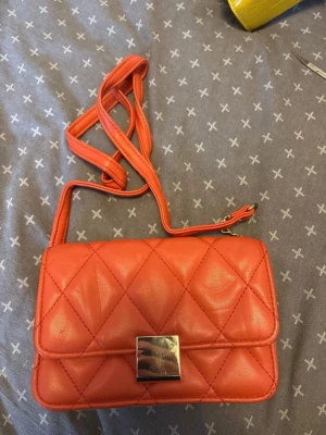 Quiltad orange axelväska - Snygg orange axelväska med quiltat mönster och justerbar rem. Väskan har en rektangulär form och stängs med ett stilrent silverfärgat spänne. Perfekt för dig som vill ha en färgklick till outfiten. Materialet är konstläder.