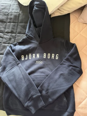 Mörkblå hoodie från Björn Borg - Snygg mörkblå hoodie från Björn Borg med vit logga och texten '1956' på bröstet. Klassisk modell med huva och magficka, perfekt för en avslappnad stil. Mjuk bomullsblandning och ribbade muddar vid ärmslut och nederkant.