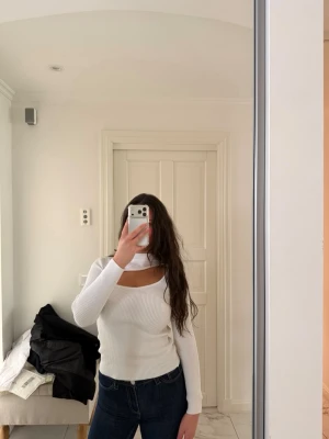 Vit ribbad topp med cut out-detalj - Trendig vit långärmad topp med ribbad struktur och snygg cut out-detalj vid halsen. Figurnära passform och hög hals ger en modern look. Perfekt att styla med jeans eller kjol för en cool outfit.
