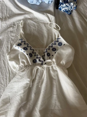Vit broderad ärmlös sommarklänning - Supersöt vit klänning med tunna axelband och blå broderade blommor vid bysten. Klänningen har v-ringning, knyt i midjan och är tillverkad i ett lätt, strukturerat bomullstyg. Perfekt för varma dagar och ger en fräsch sommarkänsla.