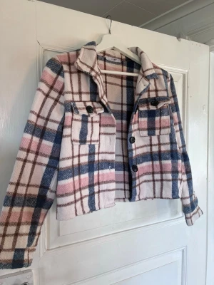 Rutig overshirt från Hailys - Trendig overshirt i mjukt material från Hailys. Jackan har rutigt mönster i rosa, blått, vitt och brunt, två bröstfickor med knappar och klassisk krage. Perfekt för lager-på-lager och en chill streetstyle-look.