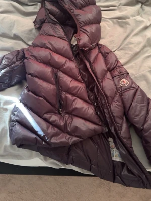 Mörklbå dunjacka från Moncler - Snygg mörkblå dunjacka från Moncler med glansig finish och huva. Jackan har snedställda dragkedjefickor och klassisk Moncler-logga på ärmen. Fodrad med dun för extra värme och har en unik serietidningsetikett på insidan.
