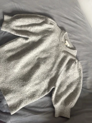 Grå stickad tröja från H&M - Mysig grå stickad tröja från H&M med puffärmar och ribbade muddar vid ärmslut och hals. Tröjan har en rak passform och är tillverkad i ett mjukt material som passar perfekt för kyligare dagar.