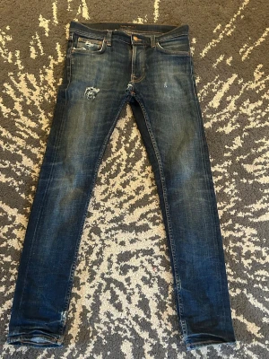 Mörkblå slim jeans från Nudie - Snygga mörkblå skinny jeans från Nudie med slitningar och lätt distressed detaljer på framsidan. Klassisk femficksmodell med läderpatch bak i midjan. Jeansen har en smal passform och är tillverkade i denim med stretch för extra komfort. Notera att de är hål i skrevet som eventuellt behövs lagas