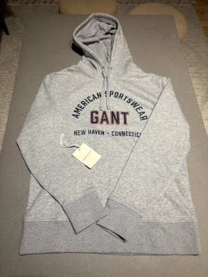 Grå hoodie från GANT med tryck - Snygg ljusgrå hoodie från GANT med klassiskt tryck på bröstet i marinblått och rött. Modellen har huva med dragsko och breda muddar vid ärmslut och nederkant. Tröjan är helt ny och kostar 1300 nypris. 