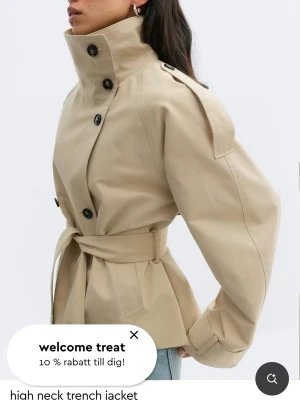 Beige trenchcoat från Gina Tricot - Stilren beige trenchcoat från Gina Tricot med dubbelknäppning, högt krage och axelklaffar. Jackan har ett avtagbart midjebälte och svarta knappar. Tillverkad i ett slätt tyg som ger en klassisk look.Jag säljer denna jacka eftersom den är helt ny och jag köpte den igår, men jag vill tyvärr inte ha den längre. Det går inte att lämna tillbaka den eftersom jag har tappat bort kvittot.