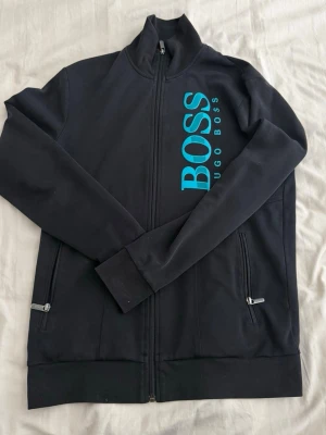 Svart zip-hoodie från Hugo Boss - Svart hoodie med dragkedja från Hugo Boss. Stor turkos logga på framsidan och två fickor med dragkedja. Bra skick och inga problem med den. 