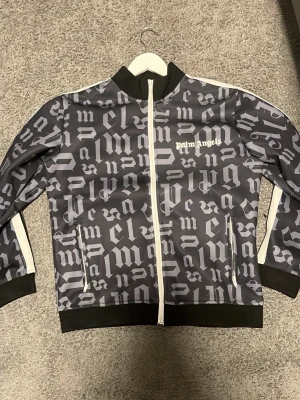 Palm Angels svart tröja med tryck - Svart tröja från Palm Angels med hel dragkedja och grått all-over-tryck. Vit logga på bröstet, vita detaljer längs ärmarna och svarta muddar. 