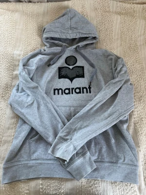 Grå Isabel Marant hoodie - (Pris går att diskutera vid snabb affär) Snygg grå hoodie från Isabel Marant med svart logga och tryck framtill. Det har lossnat en del ”ludd” från själva loggan men inget jag tänkt på när jag använt den! Klassisk modell med huva och snören samt känguruficka. Mjuk bomullsblandning som är perfekt för en chill och avslappnad stil. Storlek XL så sitter mysigt oversized! 