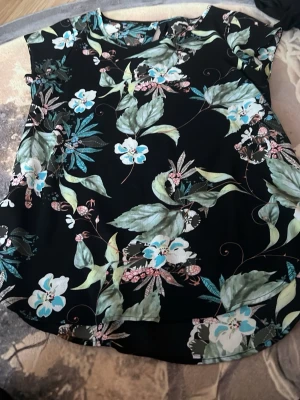 Blommig topp från ONLY - Snygg svart topp från ONLY med blommigt mönster i grönt, blått och vitt. Toppen har rund halsringning och är ärmlös, perfekt för en fräsch och trendig look. Tillverkad i lätt polyester som känns skön mot huden.