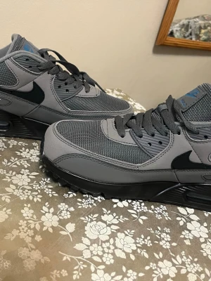 Nike Air Max 90 grå/blå sneakers - Nike Air Max 90 sneakers i grått med svarta detaljer och blå loggor. Ovandelen är i mesh och syntetmaterial för en sportig vibe. Klassisk Air Max-sula för extra komfort och stil. Perfekta för dig som gillar streetwear och vill sticka ut med en clean look.