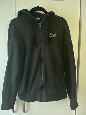 Svart zip-hoodie från EA7 Emporio Armani - Svart hoodie med dragkedja från EA7 Emporio Armani. Tröjan har huva med snören, logga på bröstet och diskreta vita ränder på ryggen. Tillverkad i mjukt bomullsmaterial och har ribbade muddar vid ärmslut och nederkant.