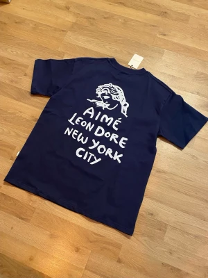 Mörkblå Aimé Leon Dore t-shirt - Snygg mörkblå t-shirt från Aimé Leon Dore med vit grafisk print och text både fram och bak. Klassisk passform med korta ärmar och rund hals. Perfekt för dig som gillar streetwear och vill sticka ut med en unik design från New York.