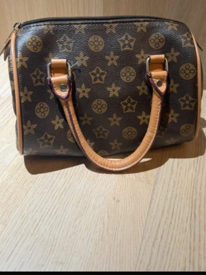 Brun handväska med monogrammönster - Snygg brun handväska med klassiskt monogrammönster i beige och guldtoner. Väskan har två korta handtag i ljust läder och dragkedja upptill. Den boxiga formen ger en trendig look och materialet är tåligt konstläder.