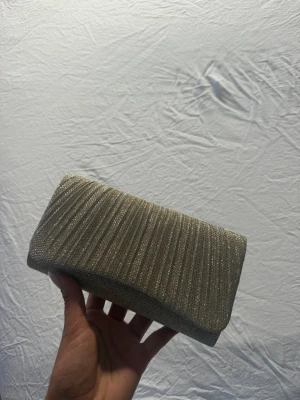 Glittrig guldig kuvertväska - Snygg kuvertväska i guldig färg med glittrigt tyg och veckad framsida. Väskan har en rektangulär form och stängs med lock. Perfekt accessoar för att lyfta din outfit med lite extra bling. Väskan är helt ny och har aldrig kommit till användning. 