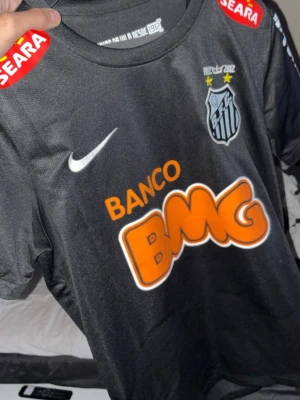 Svart Santos FC matchtröja Nike - Snygg svart matchtröja från Santos FC med tryckta loggor för BMG, SEARA och NETSHOES. Tröjan har klubbmärke på bröstet, Nike-logga och Dri-FIT-material som andas. Perfekt för fotbollsträning eller att bära som supporter. Fint skick, inga loggor som lossnat eller som är påväg att göra. Fint skick. Passar bra för dom som är mellan 175-180/82cm och väger runt 60-65kg