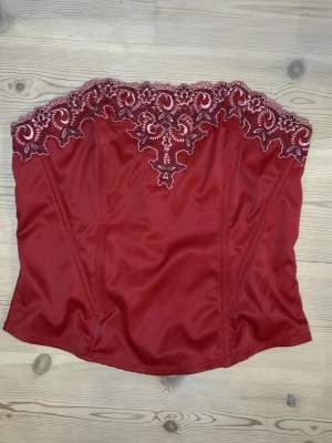 Röd korsett med spetsdetaljer - Snygg röd korsett med vacker broderad spets upptill i matchande färg. Axelbandslös modell med markerade sömmar för en figurnära passform. Perfekt för att lyfta din outfit med en touch av elegans och romantik.