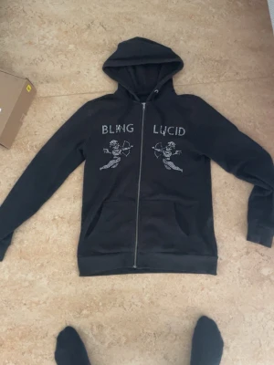 Svart hoodie från Bling Lucid - Svart hoodie med dragkedja från Bling Lucid. Framsidan har tryck med texten 'BLING LUCID' och två änglamotiv i vitt. Hoodien har huva och två fickor framtill. Perfekt för en chill och stilren look. Några pärlor saknas 