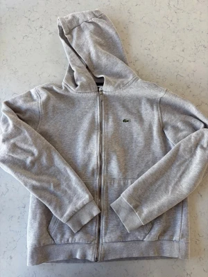Grå zip-hoodie från Lacoste - Snygg grå hoodie med dragkedja från Lacoste. Klassisk krokodillogga på bröstet och mjukt bomullsmaterial. Huva med dragsko och ribbade muddar vid ärmslut och nederkant. Inga hål eller fläckar. Bra skick. Stl 14 år, 164 cm