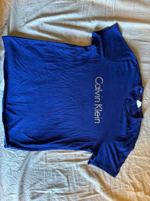 Blå Calvin Klein t-shirt - Snygg marinblå t-shirt från Calvin Klein med logga tryckt vertikalt på framsidan. Klassisk rund halsringning och korta ärmar. Tillverkad i mjuk bomull som känns skön mot huden. Perfekt för en clean och stilren look.