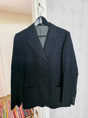 Svart klassisk kavaj med tre knappar - Stilren svart kavaj med tre knappar framtill, klassiska slag och tre fickor. Kavajen har en rak passform och är helfodrad. Perfekt för en dressad look. 100% ull. 