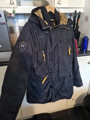 Svart parkas från Alpha Industries  - Snygg svart parkas med gula dragkedjor och detaljer. Jackan har huva med fuskpäls, flera fickor och patch på ärmen. Perfekt för kalla dagar och riktigt street. Materialet är slitstarkt syntet.