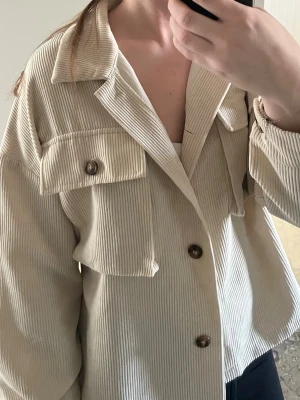 Beige jacka  - Snygg beige jacka i manchestertyg med stora bröstfickor och knappar framtill. Jackan har en avslappnad passform, klassisk krage och långa ärmar. Perfekt till sommaren eller att slänga över en t-shirt för en trendig look.