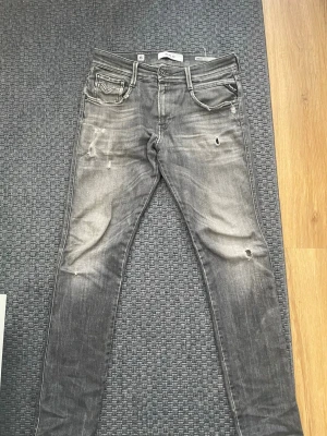Grå slitna replay Anbass  jeans   - Säljer ett par grå slim fit jeans från Replay med slitningar och distressed detaljer. Jeansen har fem fickor, dragkedja och knapp, samt Replay-logga på fickan. Perfekt för dig som gillar en tuff och trendig stil.