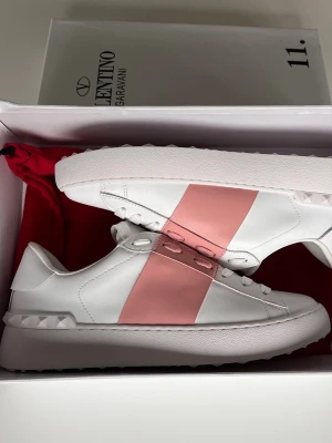 Valentino Garavani sneakers vit/rosa - Snygga sneakers från Valentino Garavani i vitt läder med bred rosa rem över vristen och coola nitar på hälen. Klassisk chunky sula och guldlogga på plösen. Perfekta för dig som vill sticka ut med stilrena detaljer och lyxig känsla.