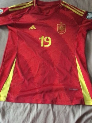 Spansk landslagströja Lamine Yamal - Röd fotbollströja från Adidas med Spaniens landslagsemblem, nummer 19 och Lamine Yamal på ryggen. Tröjan har gula detaljer och är tillverkad i lätt, ventilerande material med AEROREADY-teknologi. Perfekt för fotbollsträning eller match. Lite sliten. Tveka inte att skriva vid funderingar.