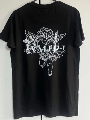 Svart AMIRI t-shirt med ängeltryck - Svart t-shirt från AMIRI med stort vitt ängeltryck och logga på ryggen samt färgstänk och logga framtill. Klassisk rund hals och korta ärmar. Tillverkad i mjuk bomull för en skön känsla och snygg passform.
