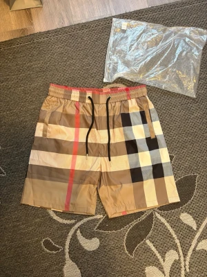 Rutig shorts från Burberry - Snygga shorts från Burberry med klassiskt rutigt mönster i beige, svart, vitt och rött. De har elastisk midja med svart snörning och sidofickor. Perfekta för en trendig och avslappnad stil.
