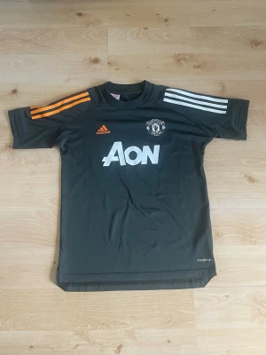 Manchester United tränings t-shirt Adidas 20/21 - Mörkgrå Manchester United tränings t-shirt från Adidas med vita och orangea ränder på axlarna. Stor vit AON-logga på bröstet och klubbmärke på vänster sida. Tillverkad i lätt och snabbtorkande material, perfekt för träning eller fotboll.